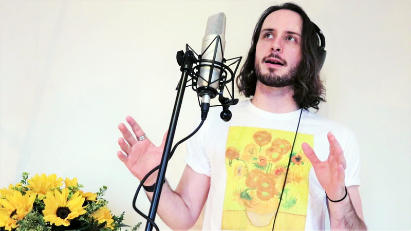 A long-haired, dark-haired man wears a white t-shirt with a print of Van Gogh's Sunflowers on the chest. He sings into a microphone which is attached to black mic stand. A bunch of sunflowers is at his right.