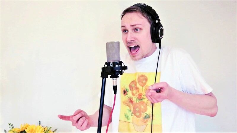A fair-haired man with a blonde beard wears black headphones and a white t-shirt with a print of Van Gogh's sunflowers on the chest. He sings into a microphone which is attached to a black mic stand with a red cable. A bunch of sunflowers stands at his right.