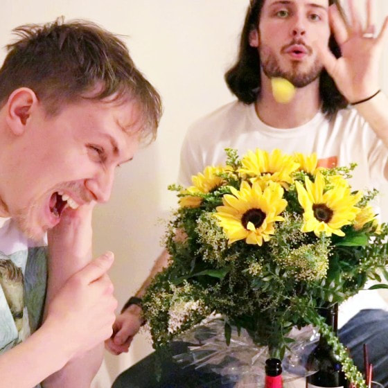 A blonde man clutches his left ear in pain. A dark-haired, bearded man blows a yellow sunflower petal into the air. They both sit next to a bunch of sunflowers.