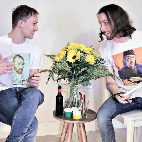 Two men in white t-shirts and blue jeans smile at one another across a bunch of sunflowers. One wears a Van Gogh portrait on his t-shirt front, the other a Paul Gauguin.